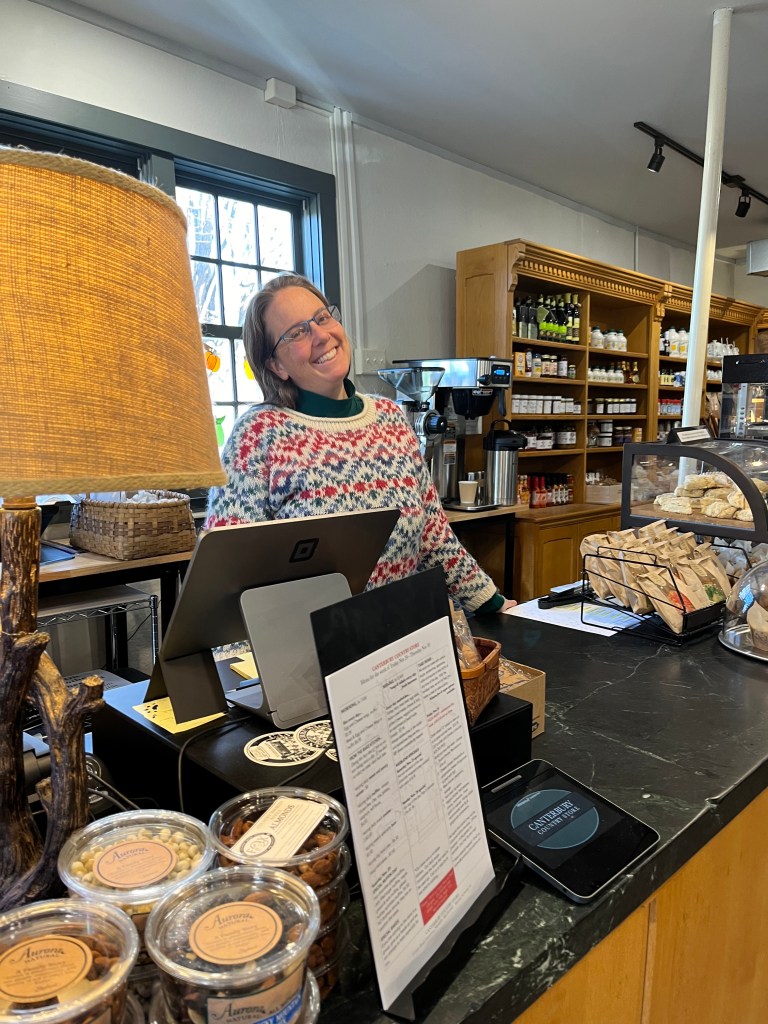 A smiling barista wearing a colorful sweater stands behind a coffee shop counter, surrounded by menus, a register, and various snacks in jars, with warm lighting and a welcoming atmosphere.