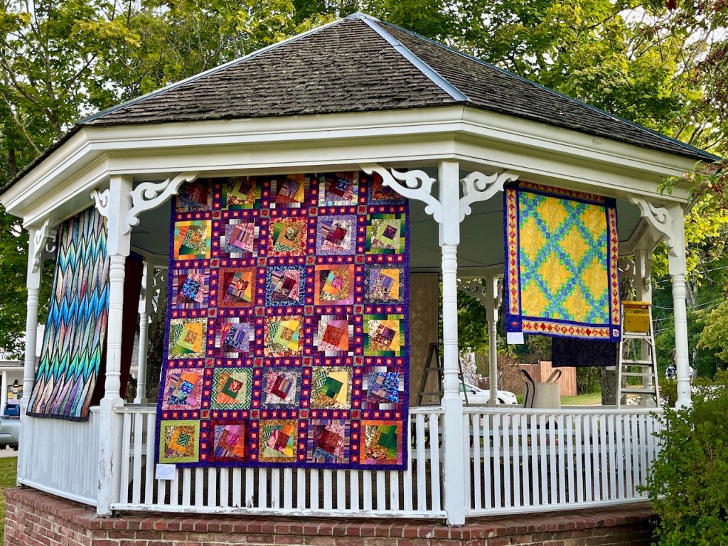 A gazebo displaying colorful quilts in various patterns, surrounded by trees and a grassy area.