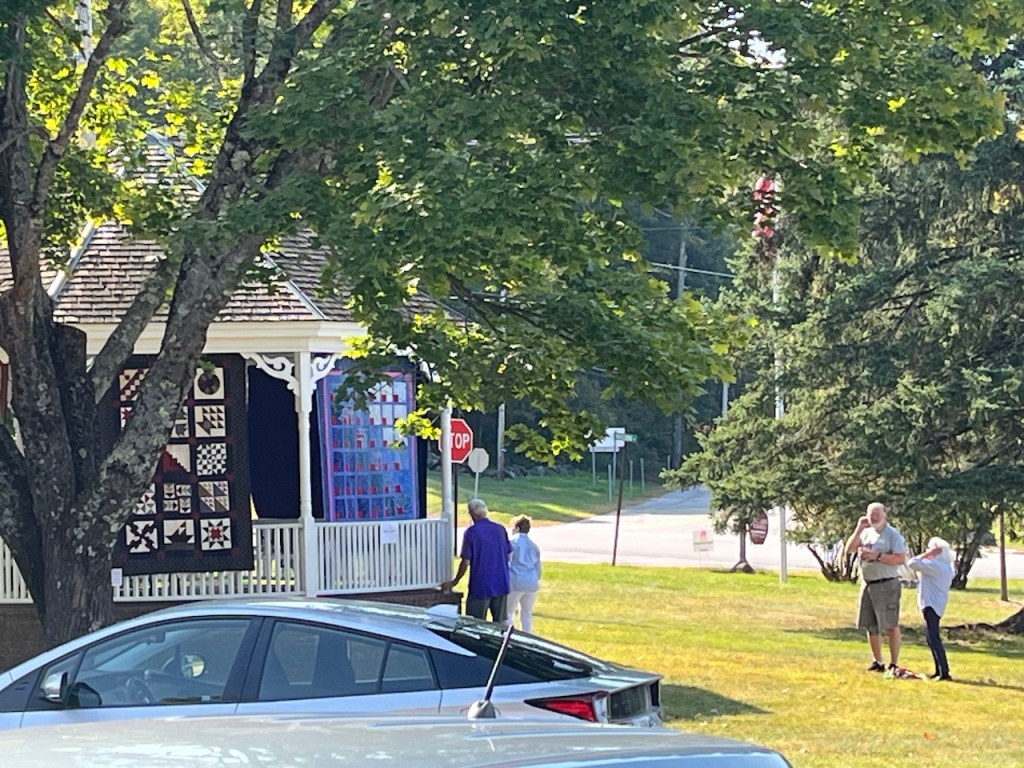 A group of four people interacting near a gazebo displaying quilt patterns, with trees and cars in the foreground.