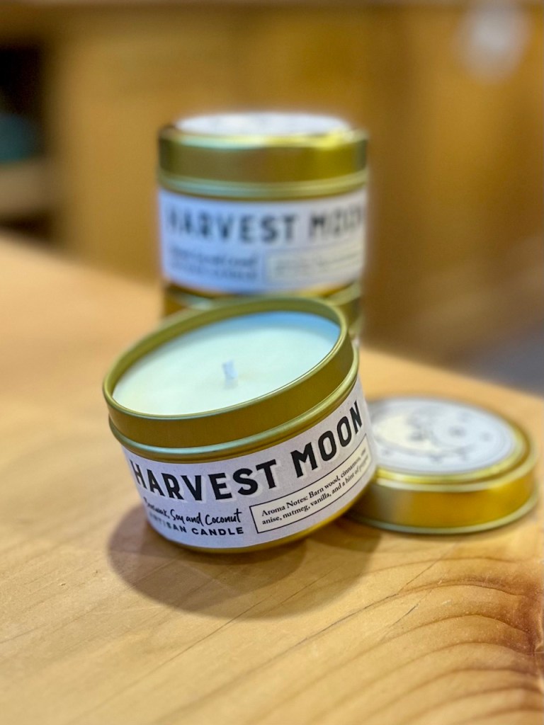 A close-up of a tin candle labeled 'Harvest Moon' with a green top, sitting on a wooden surface. Other similar candles are partially visible in the background.