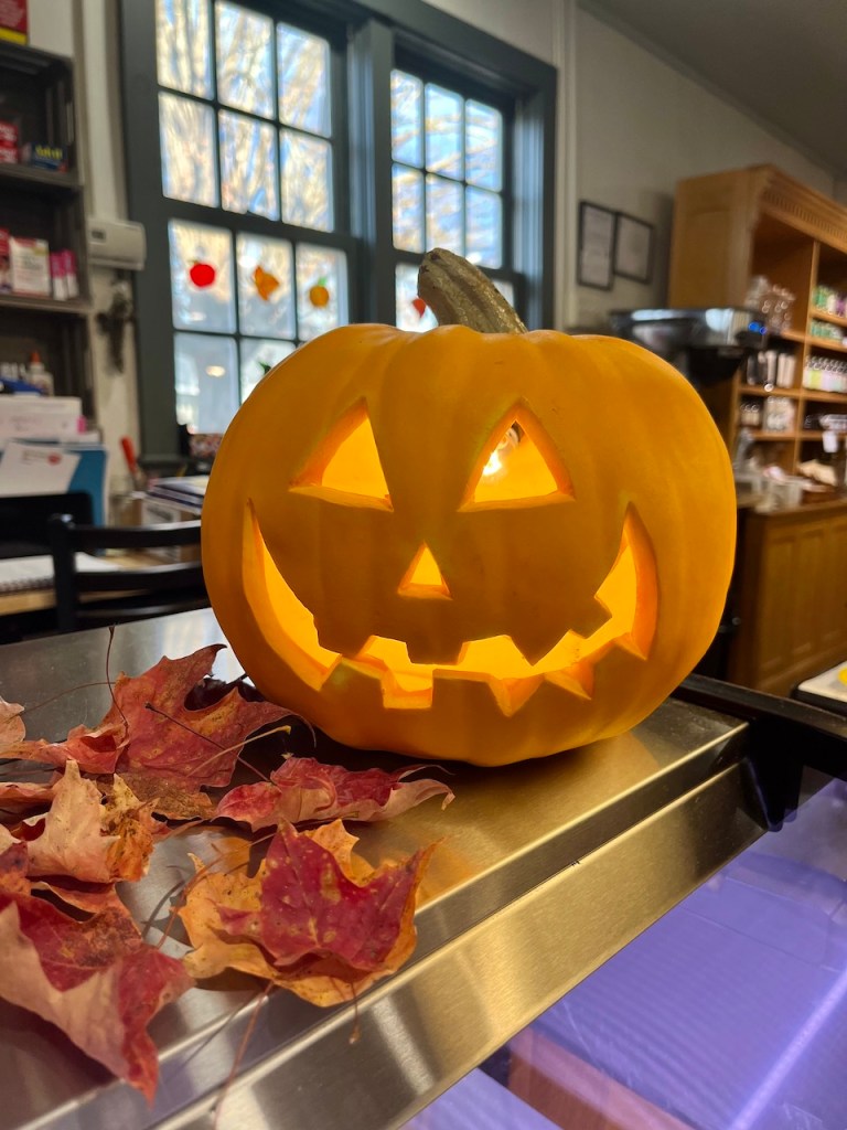 A carved pumpkin with a cheerful face, lit from within, sits on a counter next to colorful autumn leaves, showcasing a cozy fall atmosphere.