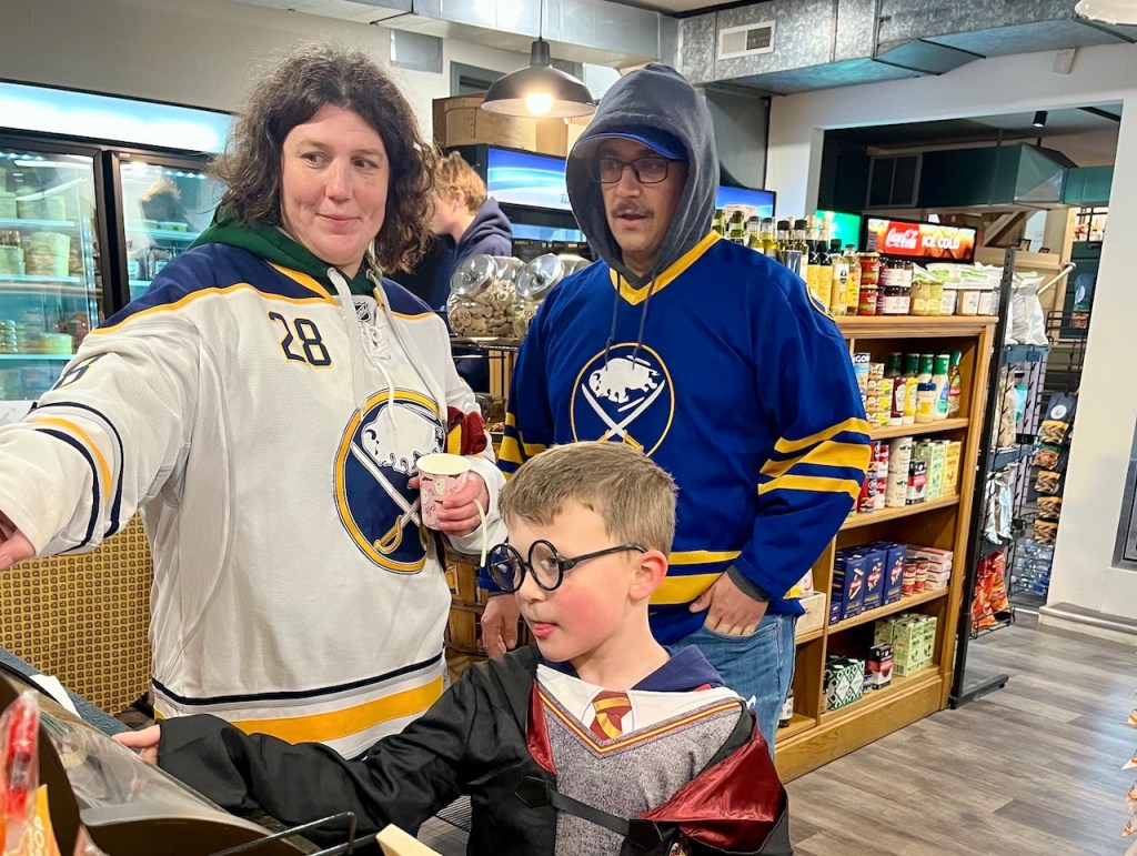A group of three people, two adults and a child, are interacting in a store. One adult wears a hockey jersey and holds a cup, while the other adult, wearing a blue hoodie, looks on. The child is dressed as a character in a wizard costume with glasses.