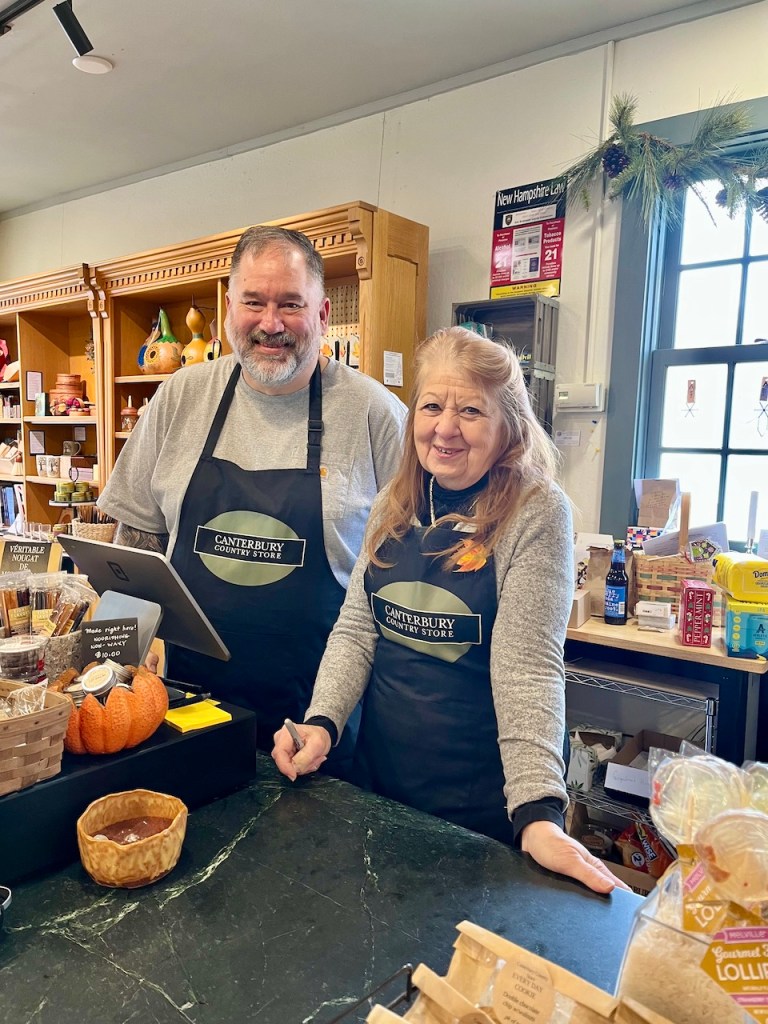 Two smiling shopkeepers standing behind the counter of a country store, wearing black aprons with the store's name, surrounded by various products and decorations.