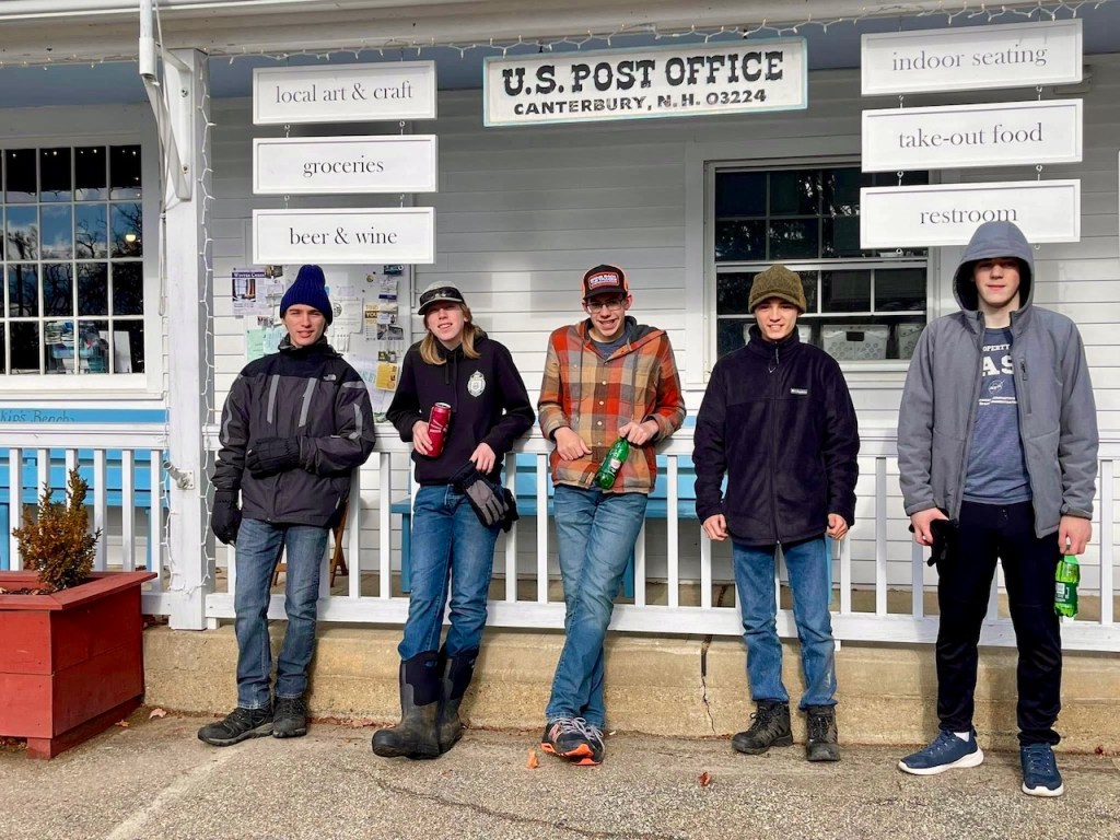 Five young individuals standing in front of the U.S. Post Office in Canterbury, New Hampshire, dressed in winter attire and holding beverages.