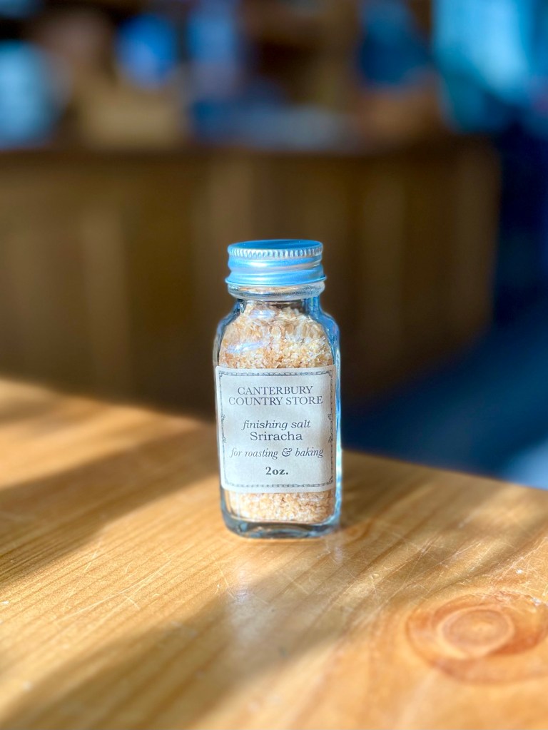 A glass jar of Sriracha finishing salt from Canterbury Country Store, placed on a wooden surface with soft lighting in the background.