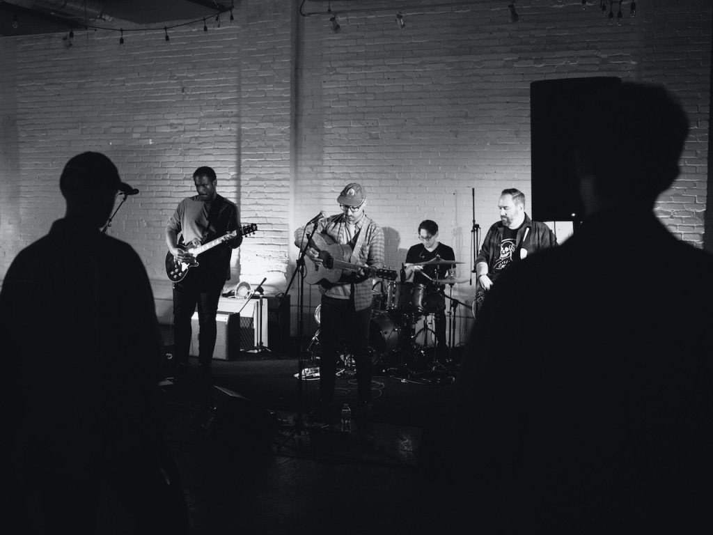 A black and white photograph of a band performing on stage, showcasing five musicians playing instruments in front of an audience. The venue features brick walls and stage lighting, creating an intimate atmosphere.