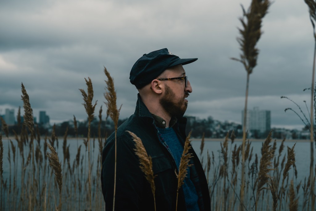 Profile of a man wearing a dark cap and glasses, standing in tall grasses by a body of water under cloudy skies.