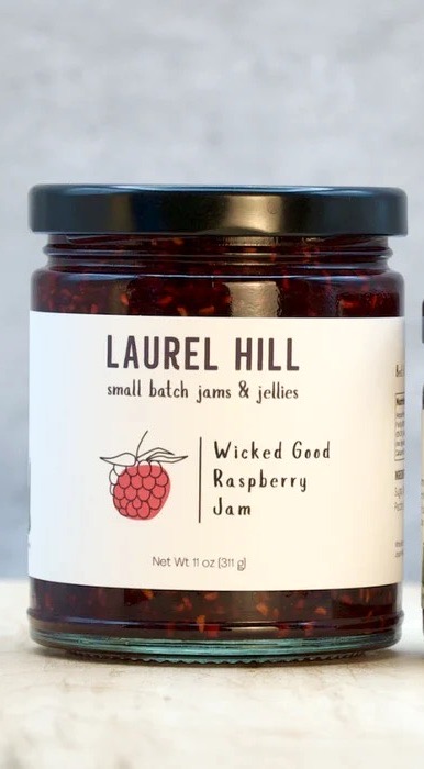 Jar of Laurel Hill Wicked Good Raspberry Jam featuring a raspberry illustration and a label detailing it as small batch jams and jellies.