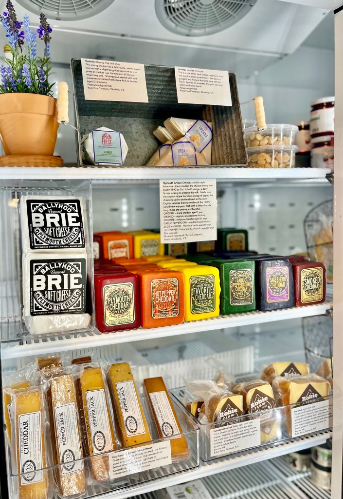 A refrigerator display filled with various cheeses, including Brie, cheddar, and flavored cheeses, alongside jars of jam and a potted plant.