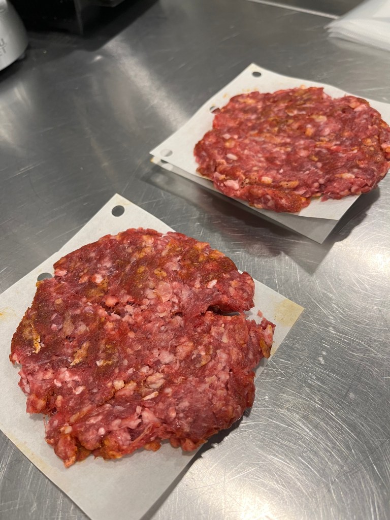 Two raw burger patties made with ground beef on parchment paper placed on a stainless steel surface.