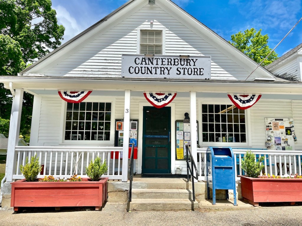 Exterior view of Canterbury Country Store, featuring a white building with a sign, decorative bunting, and flower planters on the porch.