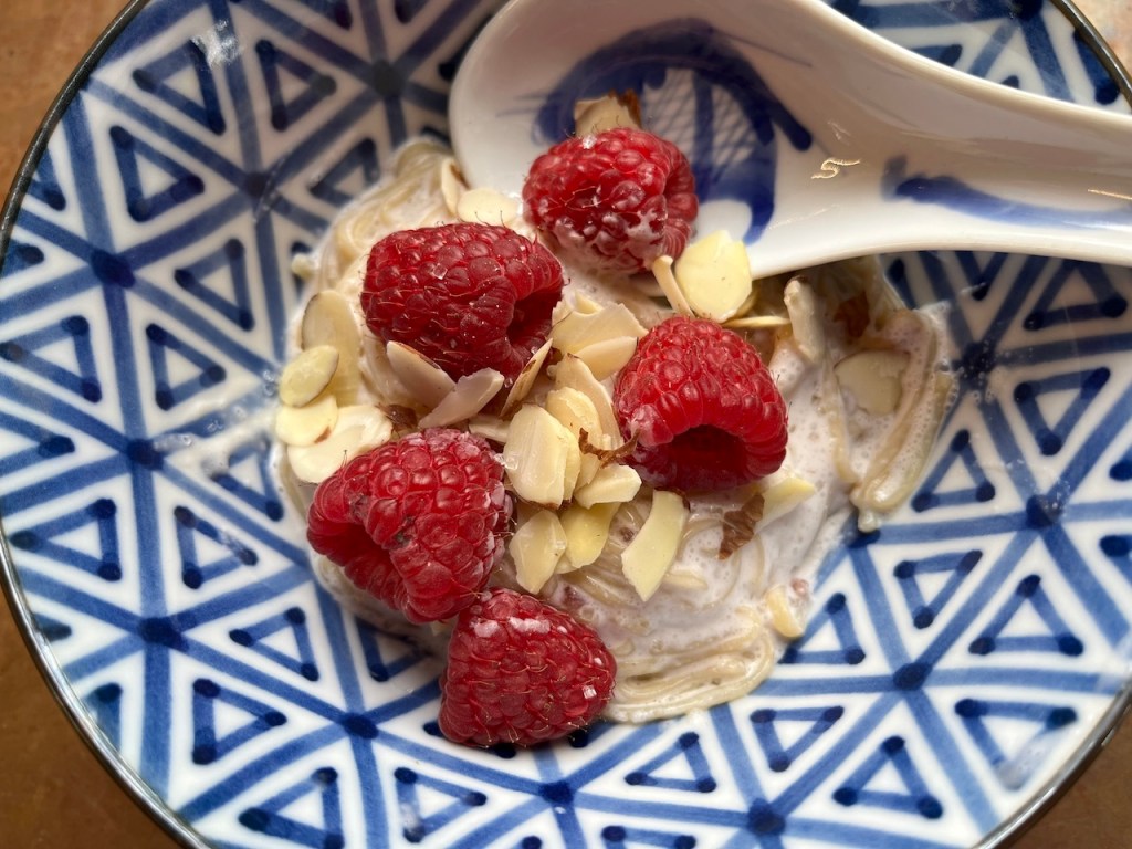A bowl of buckwheat noodles topped with fresh raspberries and slivered almonds, served with a spoon, against a decorative blue-patterned background.