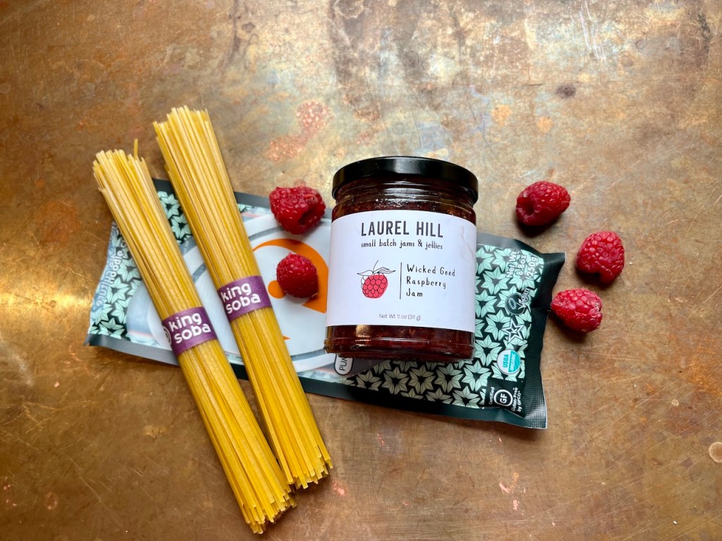 A jar of Laurel Hill Wicked Good Raspberry Jam placed on a copper surface, surrounded by buckwheat noodles, a package of King Soba noodles, and fresh raspberries.