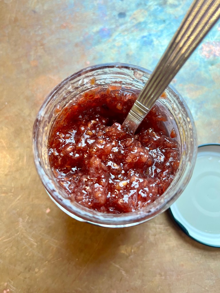 A jar filled with chunky, homemade berry jam, with a spoon resting inside, placed on a reflective surface.