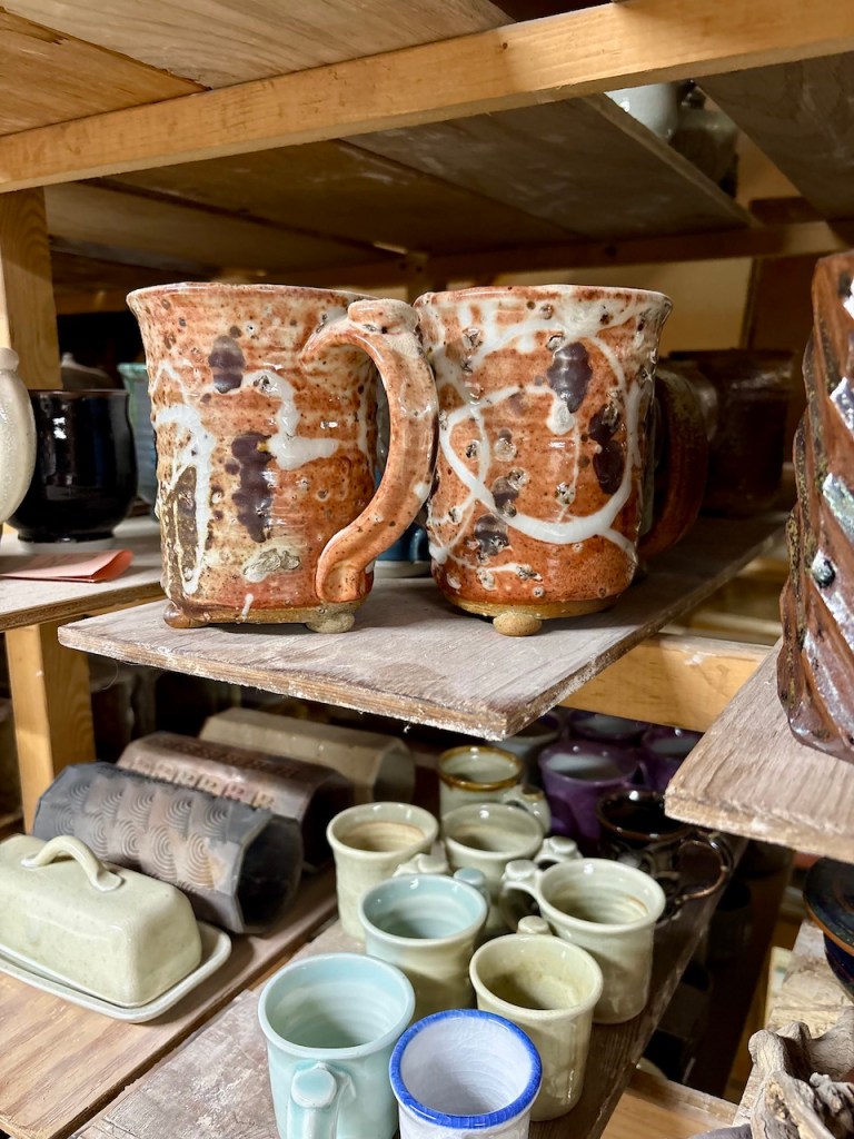 A shelf displaying various pottery items, including two rustic mugs in the foreground, featuring earthy tones and artistic glaze patterns, alongside smaller cups and dishes in different colors and styles.