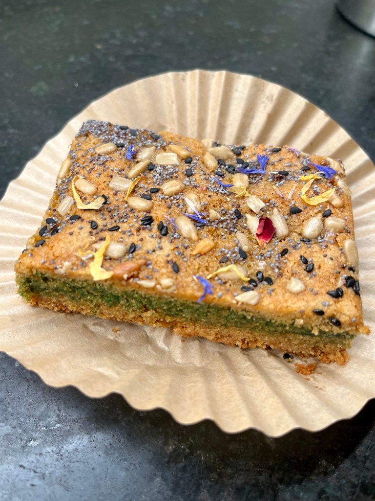 A slice of savory green herb cake topped with seeds and edible flowers, presented on a paper liner.