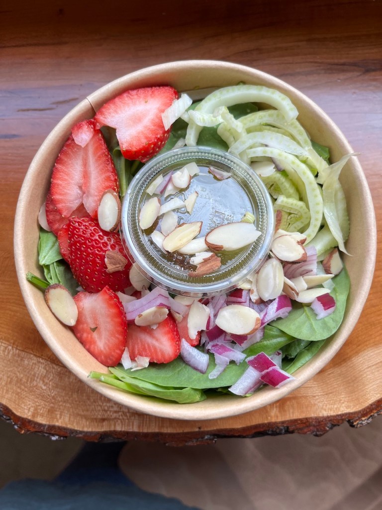 A bowl of fresh salad featuring spinach leaves, sliced strawberries, cucumber, red onion, and slivered almonds, accompanied by a small container of dressing.
