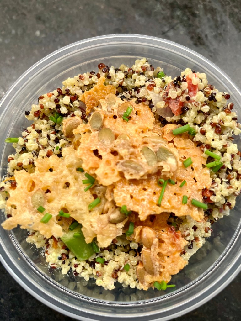A close-up view of a bowl containing a mix of quinoa, topped with crispy cheese, fresh herbs, and colorful vegetables.