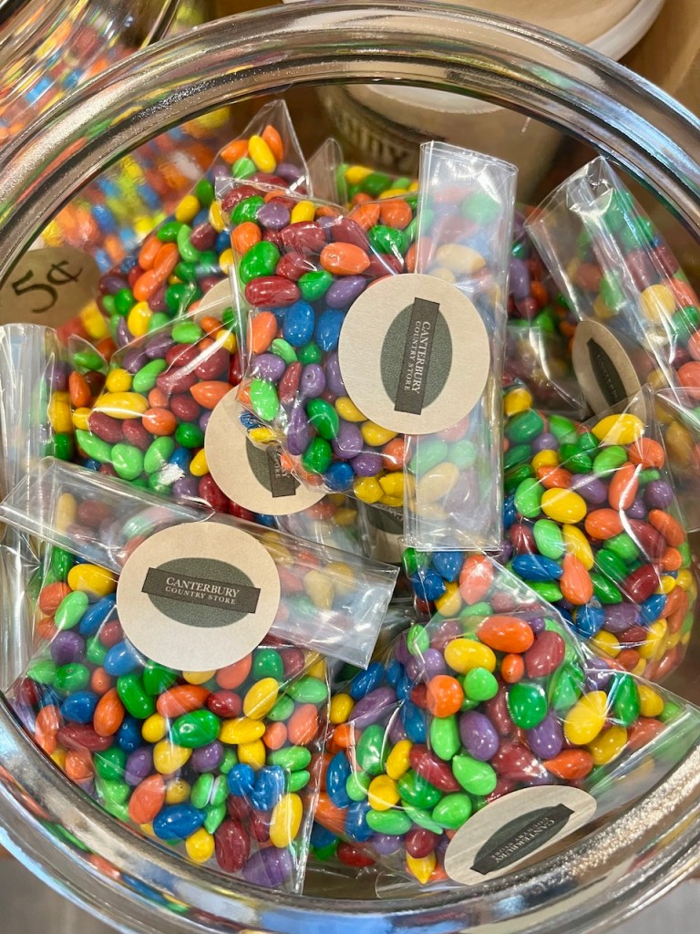 A glass jar filled with colorful candy-coated chocolates, some in small clear bags labeled with 'Canterbury Country Store'.