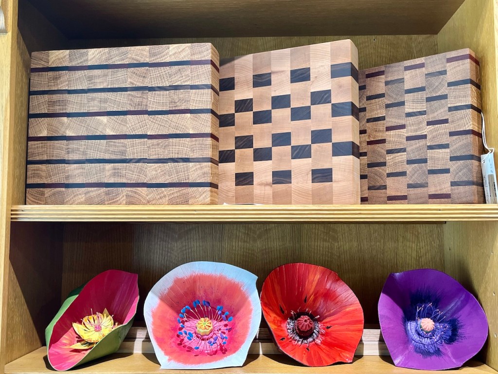 A display of wooden products including cutting boards with various patterns and colorful crafted flowers made from paper or similar materials, arranged on a wooden shelf.