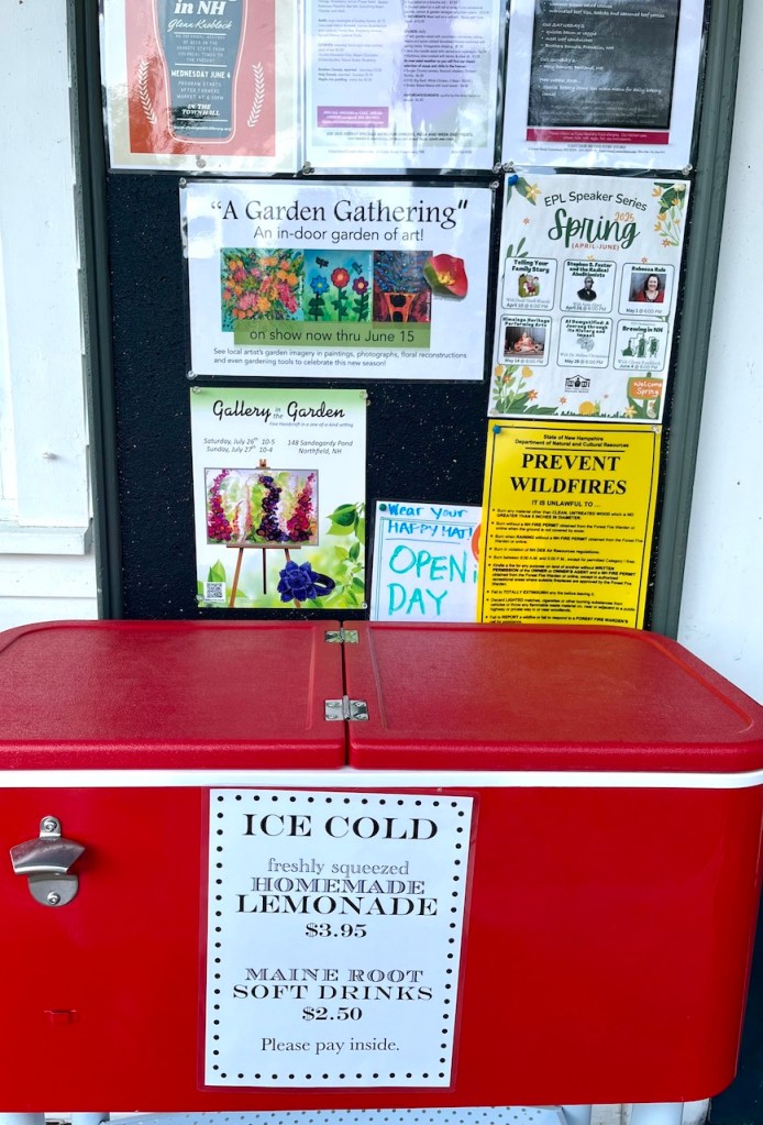 A red cooler displaying a sign for ice-cold homemade lemonade and Maine Root soft drinks, with pricing information, positioned against a bulletin board filled with flyers and announcements.