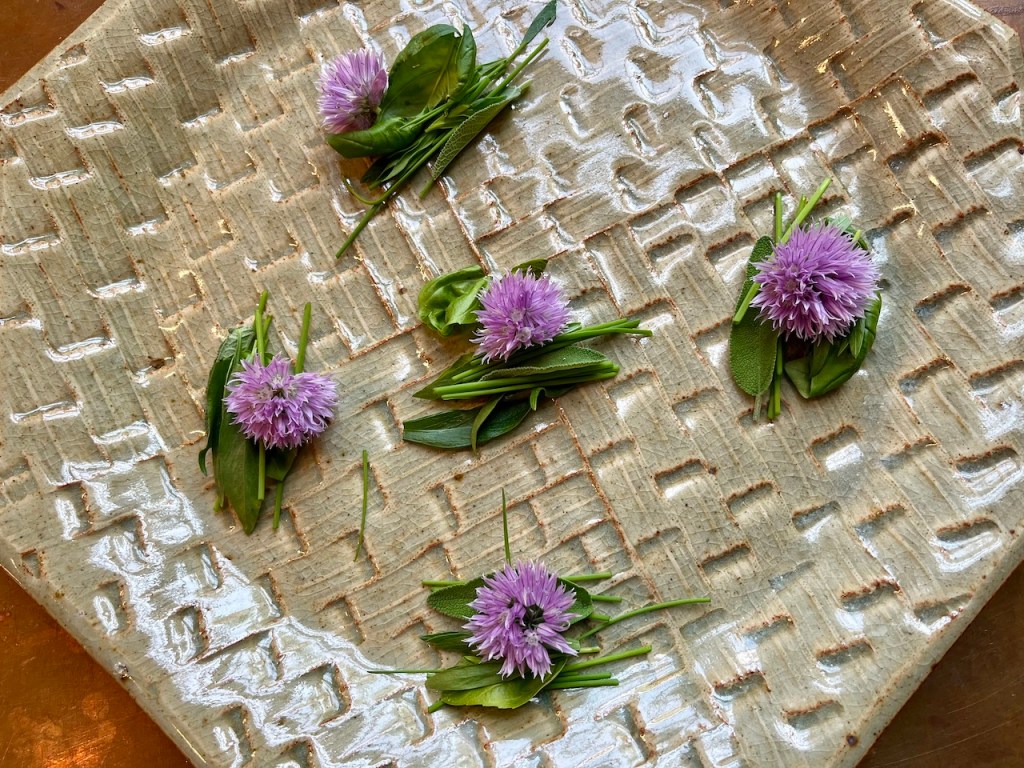 A ceramic plate decorated with arranged bundles of chive blossoms and various green herbs, such as sage and tarragon.