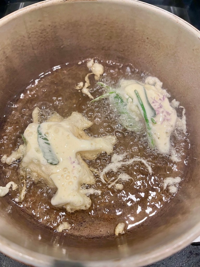Crispy herb fritters frying in oil, with bubbles forming around the batter in a pot.