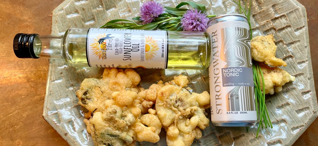 A serving platter featuring crispy herb fritters, a bottle of sunflower oil, and a can of Nordic tonic, surrounded by fresh chives and chive blossoms.