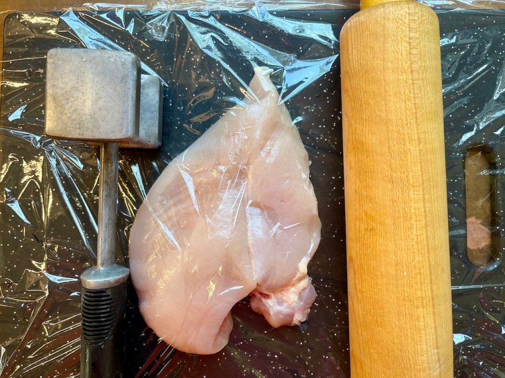 Raw chicken breast covered with plastic wrap on a cutting board, alongside a meat mallet and a rolling pin.