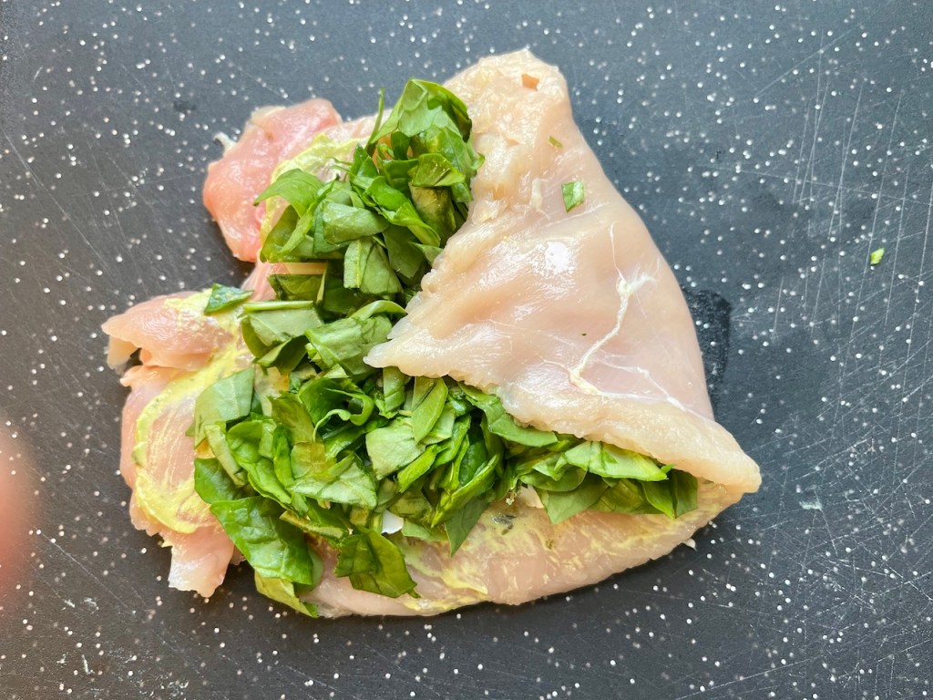 Flattened chicken breast with Dijon mustard and fresh spinach filling on a cutting board.