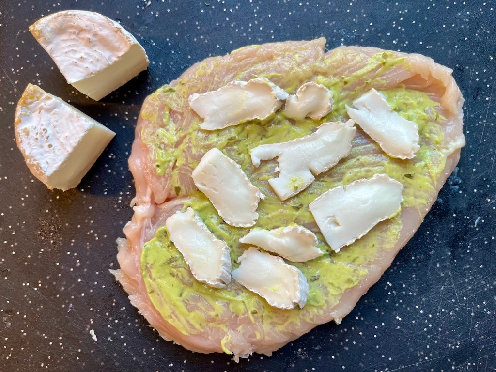 Raw chicken breast spread with tarragon mustard and slices of Brie cheese, with additional cheese pieces on the side.