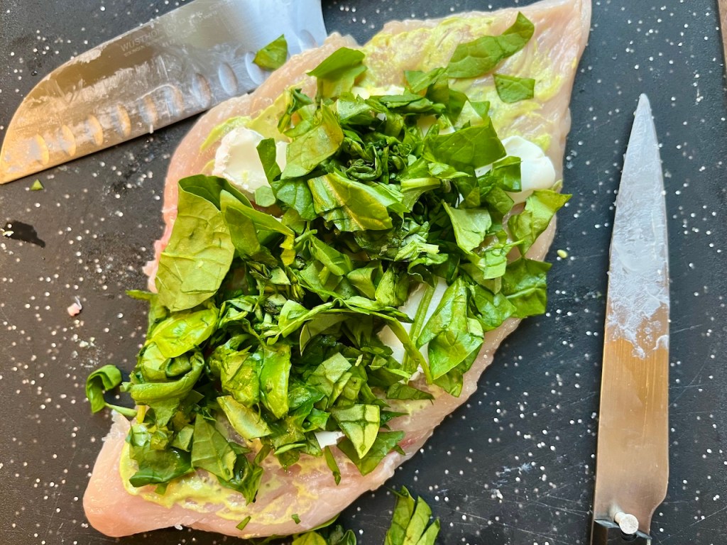 A cutting board with a flattened chicken breast topped with fresh chopped spinach and cheese, along with a knife resting beside it.
