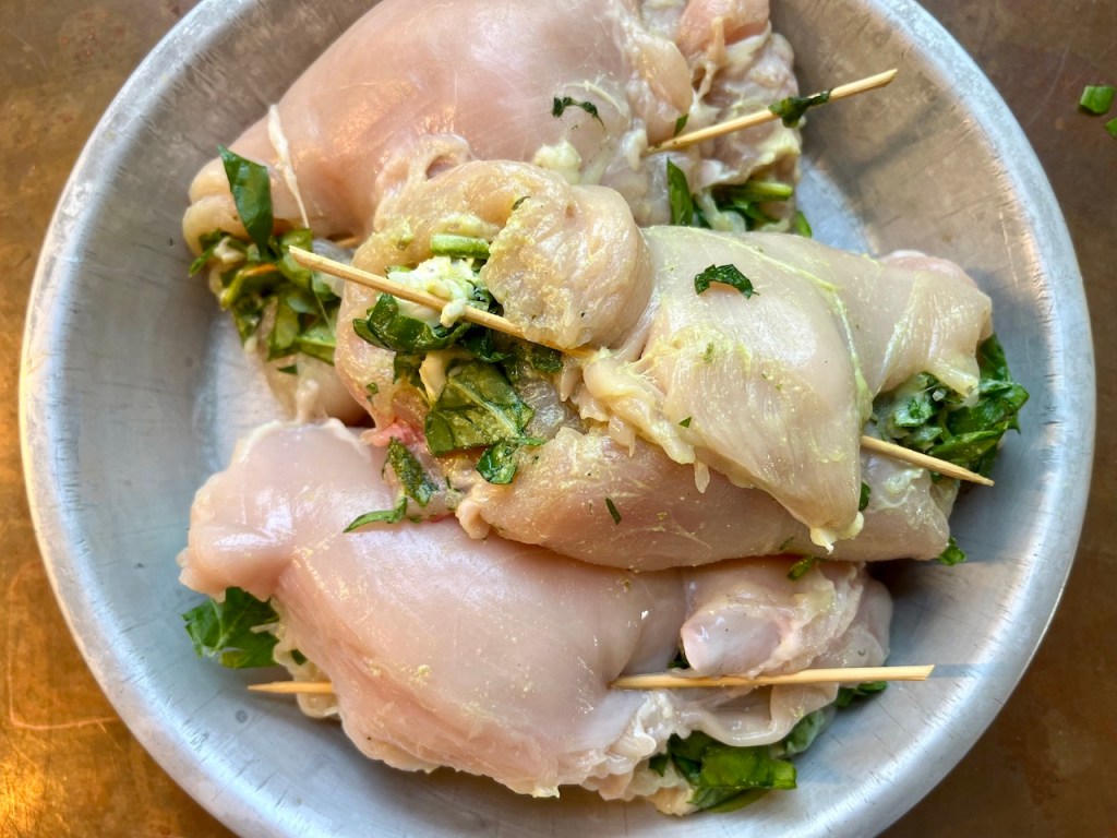 Raw chicken breasts stuffed with spinach and mustard, secured with bamboo skewers, in a round metal dish.