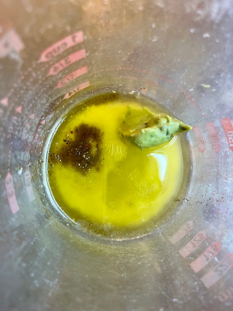 A close-up view of a measuring cup containing olive oil, mustard, and seasoning with a sprig of tarragon.