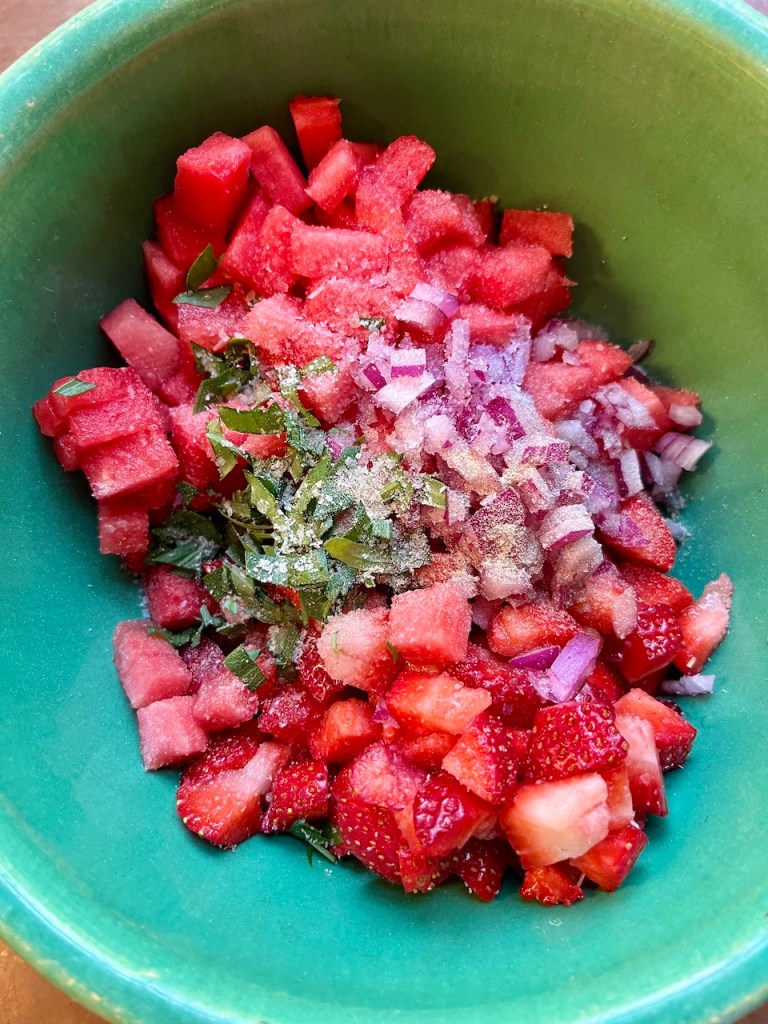 A bowl filled with diced watermelon and strawberries, minced red onion, chopped herbs, and seasoning, ready for salsa preparation.