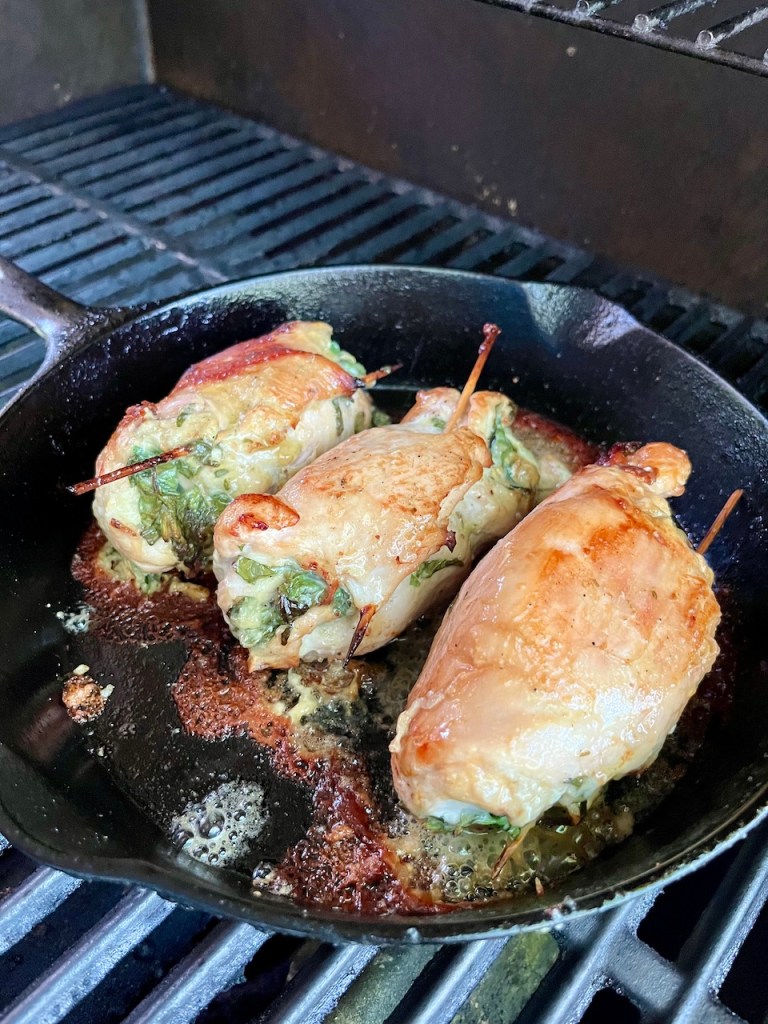Three rolled chicken breasts cooking in a cast iron skillet on a grill, garnished with herbs.