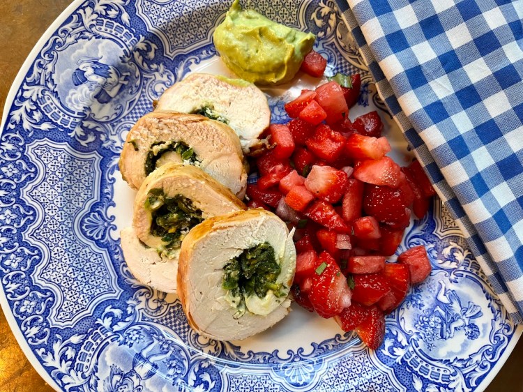 A plate featuring sliced rolled chicken breasts stuffed with spinach and cheese, accompanied by a fresh strawberry-watermelon salsa and a dollop of guacamole, set against a blue and white patterned plate with a checkered napkin.
