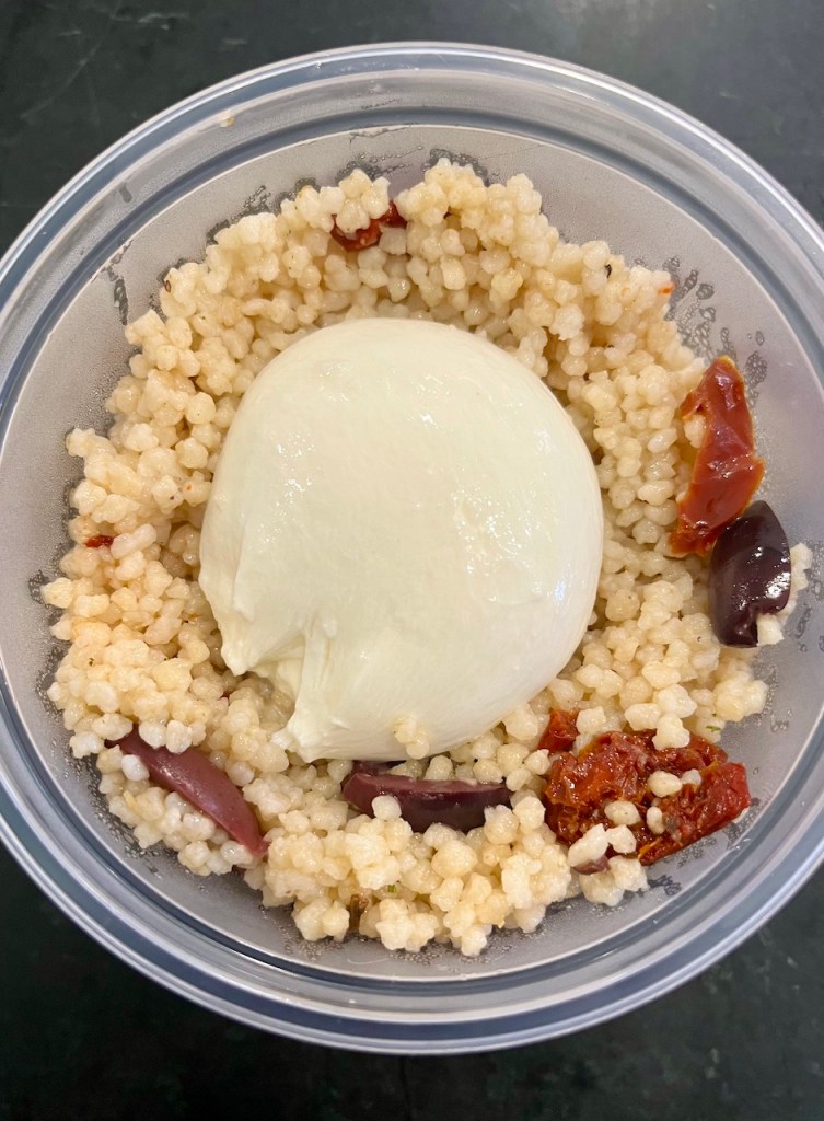 A bowl of couscous topped with a soft cheese ball and garnished with sun-dried tomatoes and olives.