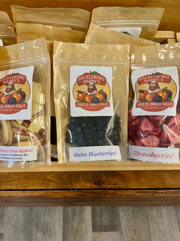 Close-up of various packages of freeze-dried fruit displayed in a wooden store shelf, including Honey Crisp Apples, Maine Blueberries, and Strawberries, all labeled with branding.
