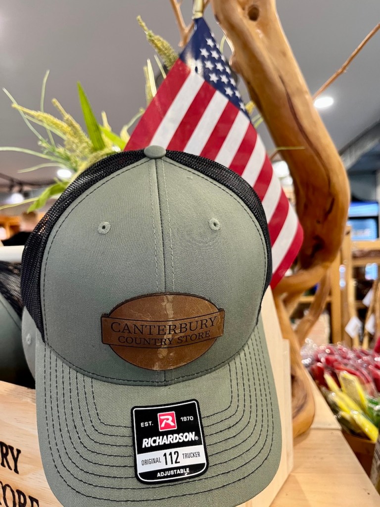 A green trucker hat with a brown leather patch labeled 'Canterbury Country Store' on the front, displayed alongside a small American flag and various rustic items in a store setting.