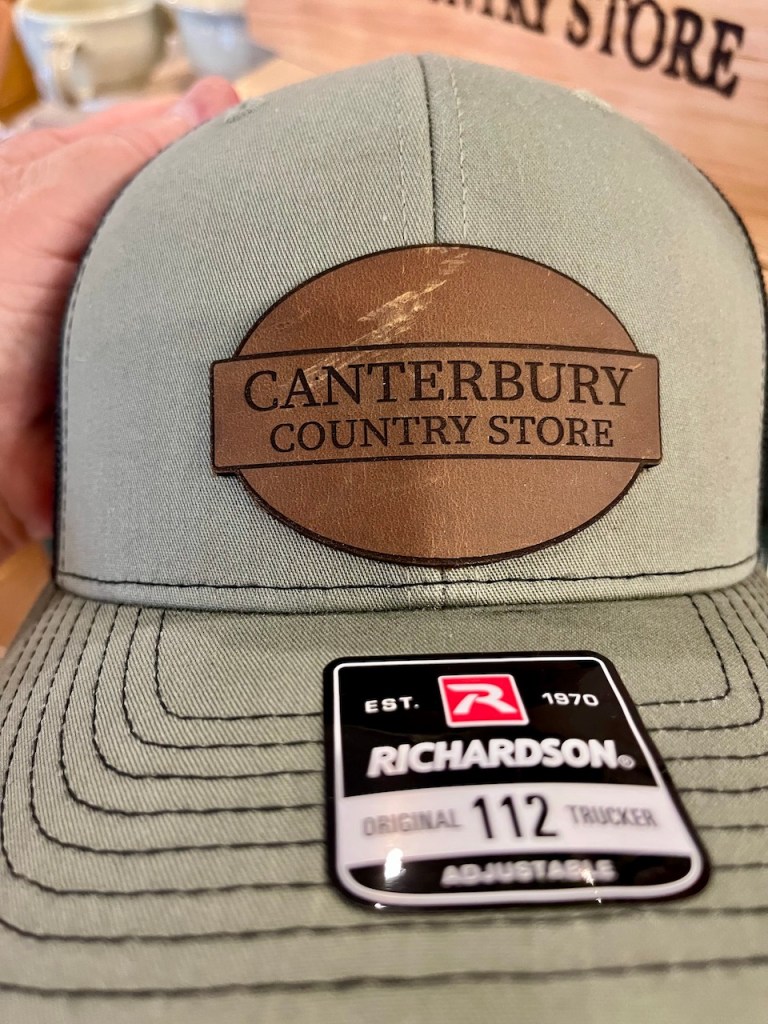 Close-up of a baseball cap with a leather patch featuring the text 'CANTERBURY COUNTRY STORE' and a tag indicating it is a Richardson Original 112 Trucker cap.