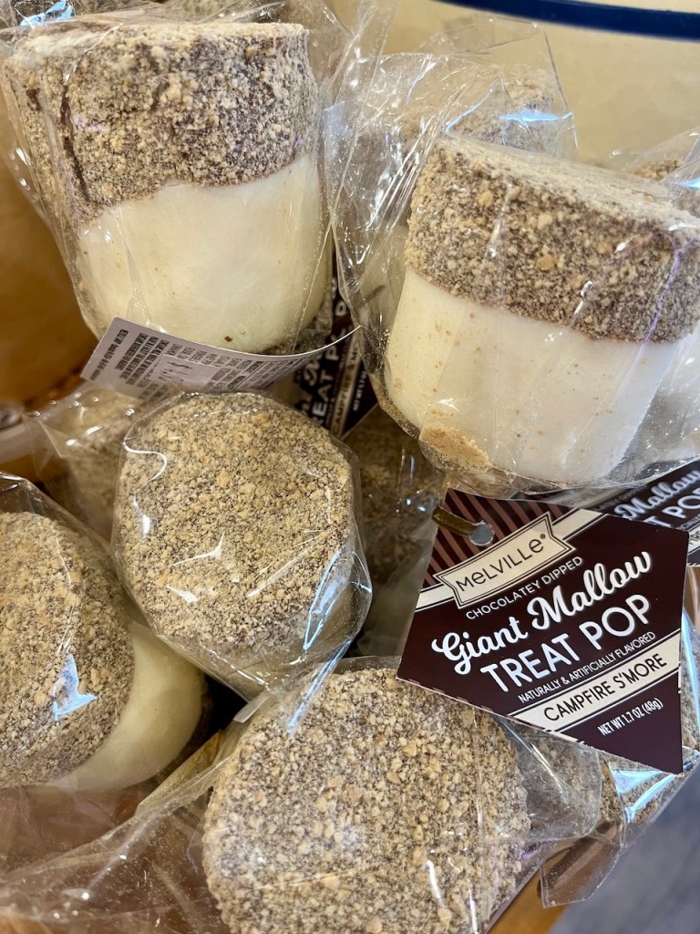 Close-up of packaged Giant Mallow treat pops, chocolate-dipped marshmallows coated in graham cracker crumbs, displayed on a shelf.