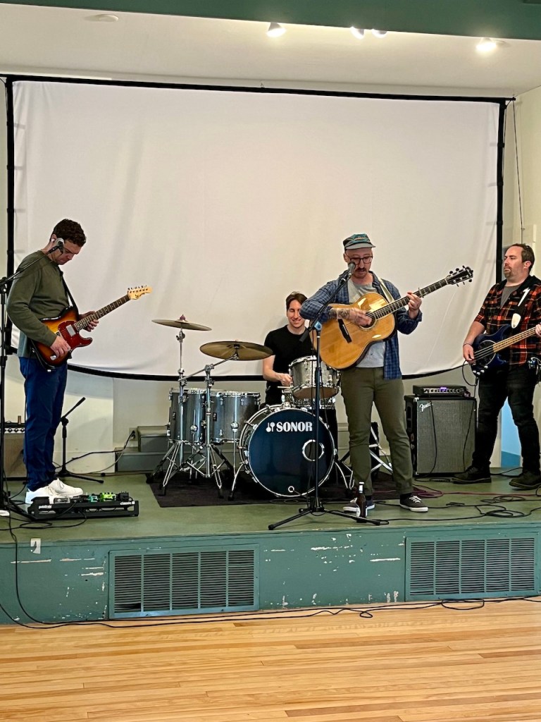 A live music performance featuring four musicians on stage, with a guitar, drums, and vocals, in a cozy venue.