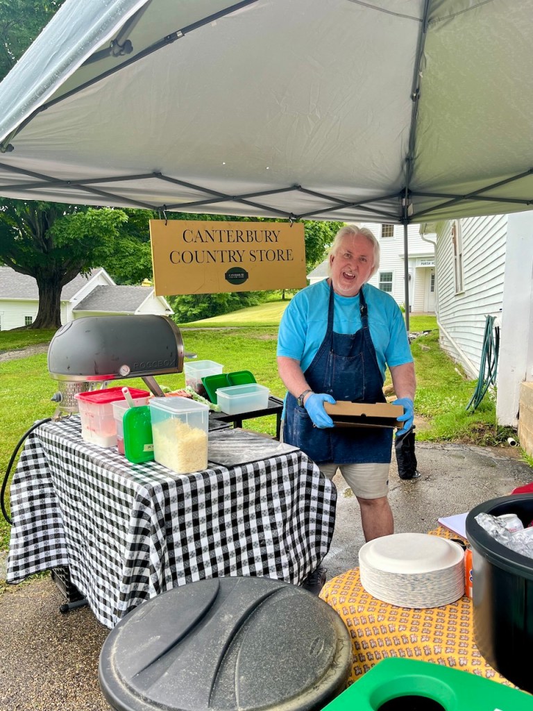 A smiling man wearing gloves stands under a tent at the Canterbury Country Store, holding a take-out box. The table is covered with a checkered cloth and has various food items and containers.
