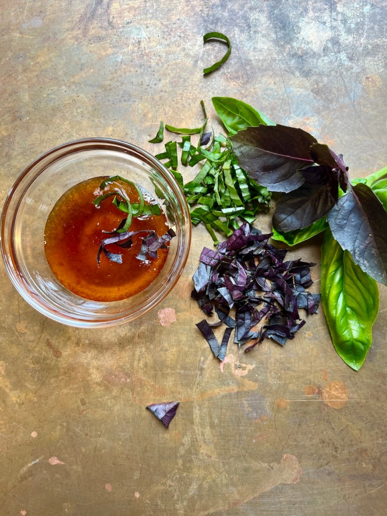 A small glass bowl filled with a mixture of honey and finely chopped basil leaves, surrounded by additional basil leaves on a textured surface.