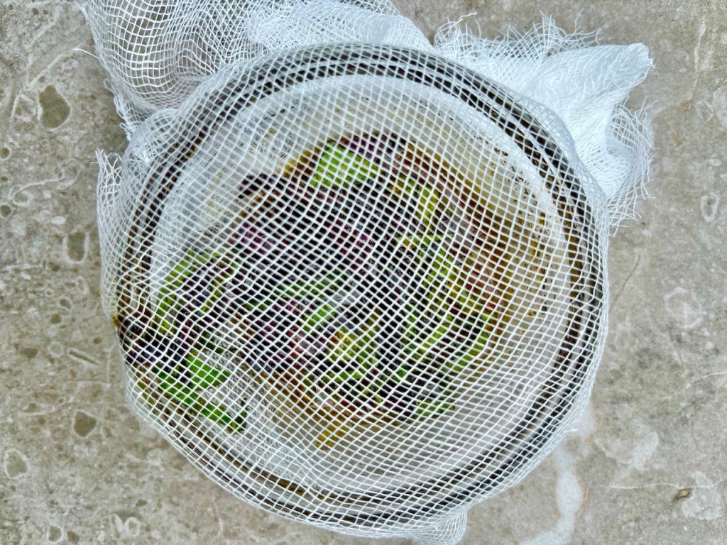 A bowl with a cheesecloth covering containing basil and honey, set on a textured surface.