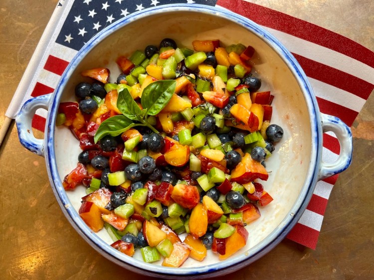 A bowl of red, white, and blue fruit salad featuring diced stone fruit, blueberries, and chopped celery, garnished with a fresh basil leaf, on a wooden surface alongside a small American flag.