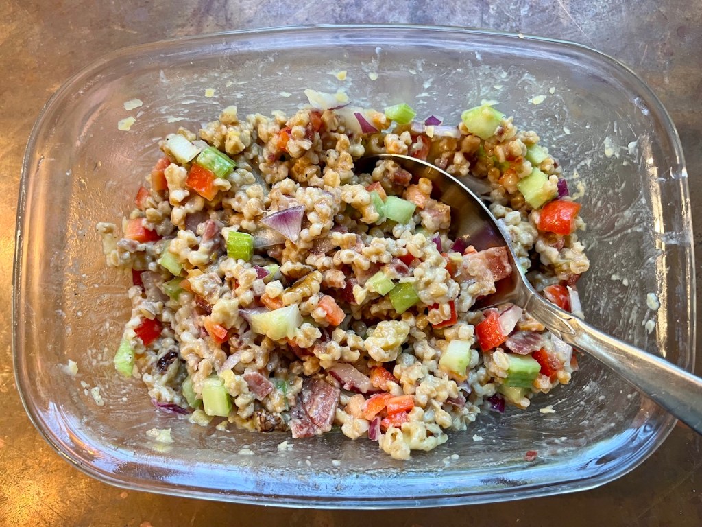 A glass bowl filled with a colorful grain salad, featuring chopped vegetables like celery, red onion, and red sweet pepper, mixed with grains and a creamy dressing.