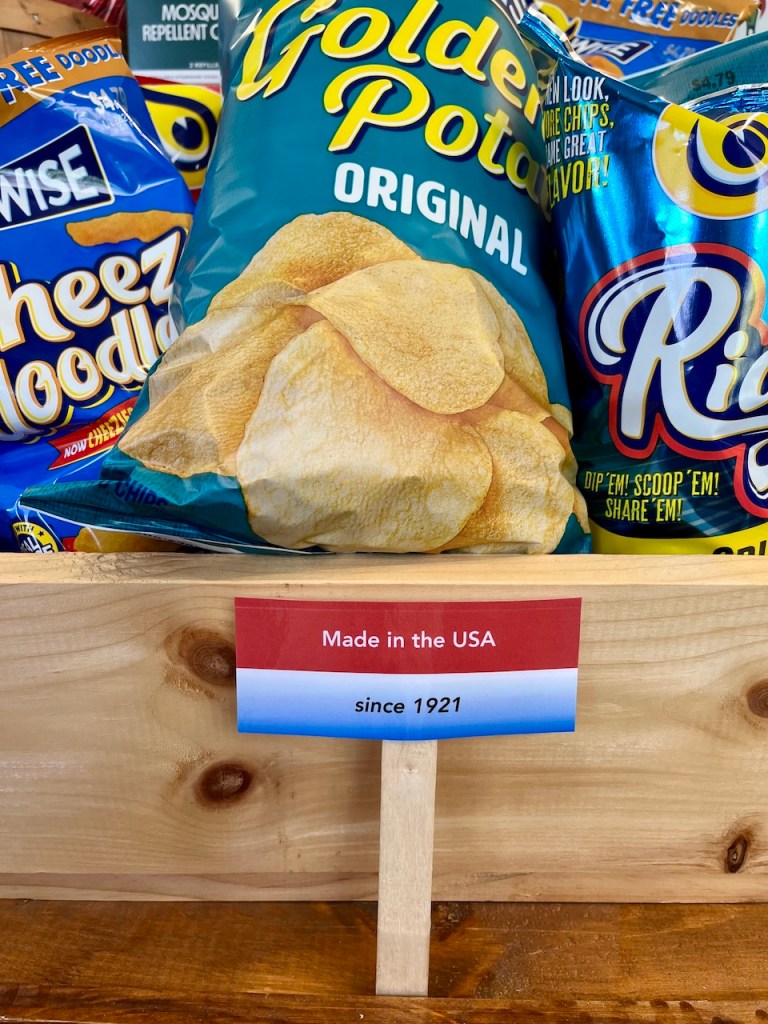 Display of snack bags including 'Golden Potato Original' chips and 'Cheez Doodles' with a sign stating 'Made in the USA since 1921' on a wooden shelf.