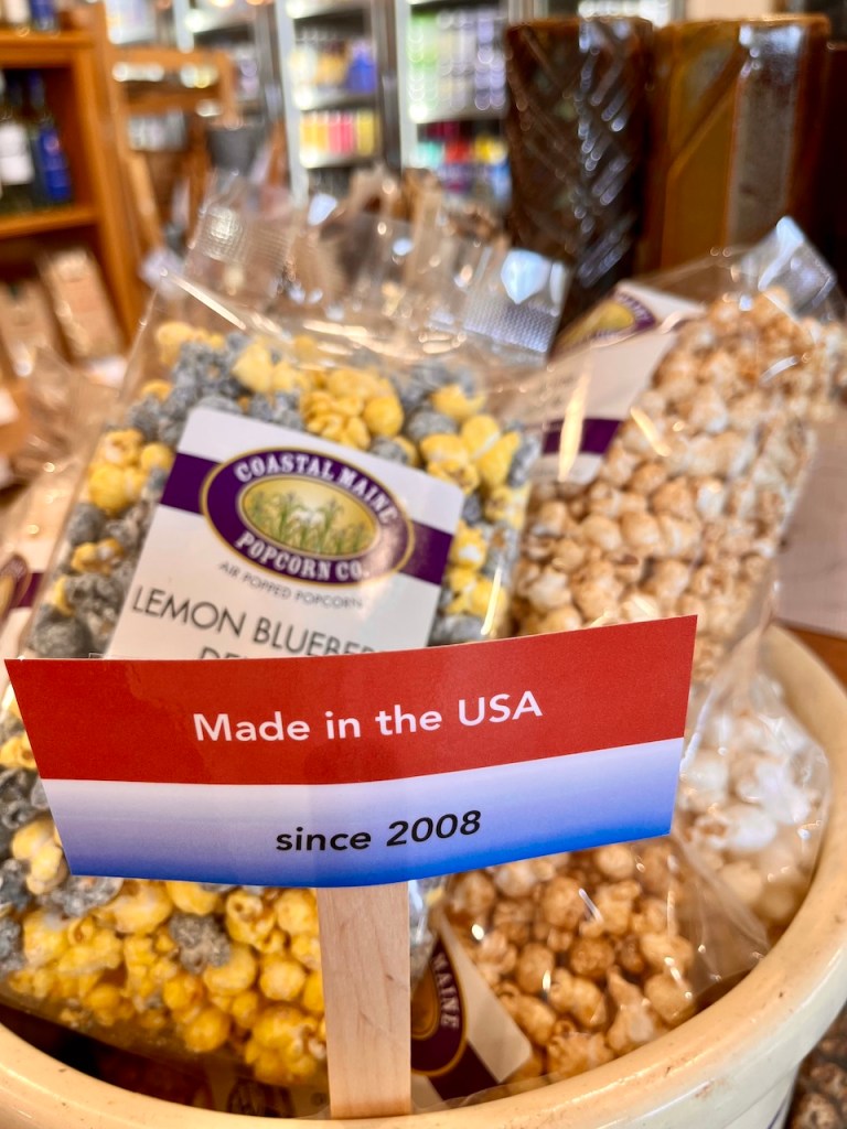 A display of bags of popcorn in a store, featuring a sign that says 'Made in the USA since 2008'. The popcorn is a mix of yellow and blue flavors, with an assortment of containers visible in the background.